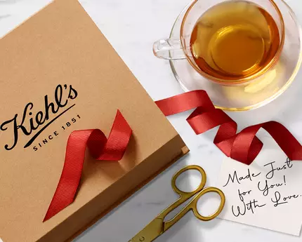 Кутия с лента с логото на Kiehl's, чаша чай и поздравителна картичка на мраморна маса.