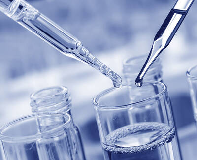 Two pipettes are positioned above a glass container filled with a clear liquid, with one pipette releasing a drop. The overall color tone is blue.