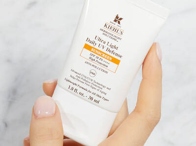 A hand holding a tube of Kiehl's Ultra Light Daily UV Defense sunscreen with SPF 50, against a light marble background.