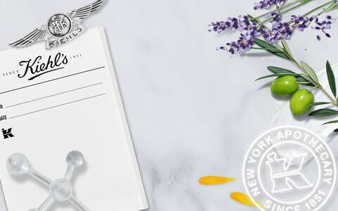 Alt text: A vintage black and white photo of Kiehl's pharmacy, featuring staff members in white coats, alongside a modern marble background decorated with lavender, olive leaves, and the Kiehl's logo, emphasizing the brand's long history since 1851.