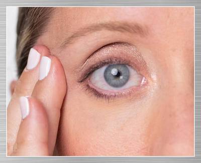 A close-up of a woman's eye, showing her eyelid and skin texture, with her fingers gently touching the outer corner of her eye. She has a subtle makeup look.