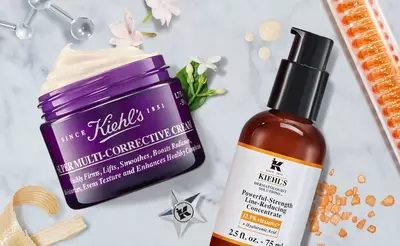 A flat lay of Kiehl's skincare products, featuring a purple jar of Super Multi-Corrective Cream and an orange bottle of Powerful-Strength Line-Reducing Concentrate, surrounded by decorative elements and textures.