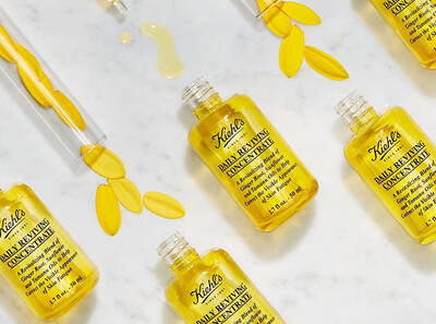 Флакони с концентрат за лице Kiehl's, разположени на мраморна повърхност, с цветя и капки масло.