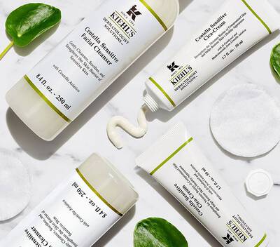 Продукти за грижа за чувствителна кожа от Kiehl's: почистващ гел и крем на бяла повърхност с памучни дискове.