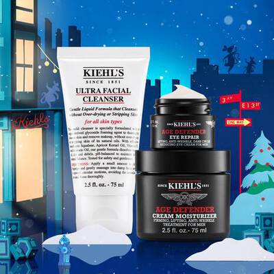 A festive scene featuring Kiehl's skincare products: a tube of Ultra Facial Cleanser, a jar of Age Defender Eye Repair, and a jar of Age Defender Cream Moisturizer, set against a wintry backdrop with holiday decorations.