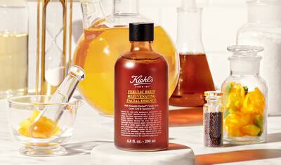 A bottle of Kiehl's Ferulic Brew Rejuvenating Facial Essence is placed on a marble surface, surrounded by various lab glassware filled with colorful liquids and ingredients, including yellow flowers and seeds.