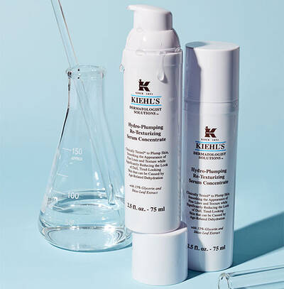 Бяло шише с серум Kiehl's на син фон, комбинирано с лабораторна стъклена посуда.