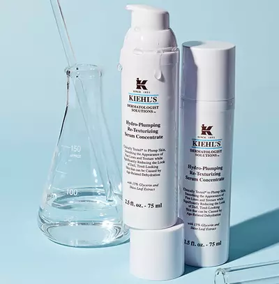 Бяло шише с серум Kiehl's на син фон, комбинирано с лабораторна стъклена посуда.