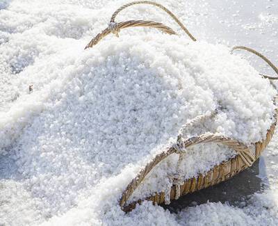 A woven basket filled with large grains of white salt, set against a sandy background.