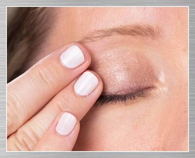 A close-up of a woman’s eye with a finger gently touching her eyelid, showcasing a subtle eyeshadow look. The nails are painted white.