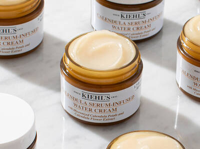 Крем с екстракт от невен и серум в стъклени бурканчета на Kiehl's, разположени на мраморна повърхност.