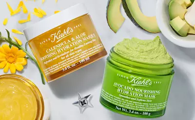 Two jars of Kiehl's skincare masks are displayed on a textured surface. The first, labeled "Calendula & Aloe Soothing Hydration Mask," is a cream-colored jar; the second, "Avocado Nourishing Hydration Mask," is a green jar. Surrounding the jars are petals, avocado slices, and a small bowl of gel