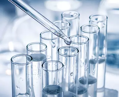 A pipette is dripping liquid into a row of clear test tubes, each containing a small amount of liquid. The background features blurred laboratory equipment.